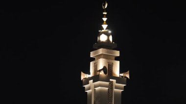 Gece vakti aydınlatılmış Cami Minaresi. Güzel aydınlatılmış bir cami minaresi gece gökyüzüne karşı duruyor. Karmaşık tasarım ve parlak ışıklandırma İslam 'ın mimari güzelliğini vurguluyor.
