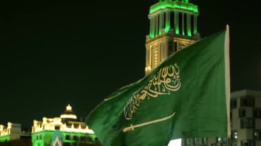 Ulusal Bayram kutlamalarında Suudi Bayrağı Gece Dalgalanıyor. Suudi Arabistan bayrağının yakın çekimleri Ulusal Bayram 'ı simgeleyen yeşil ışıklarla süslenmiş aydınlanmış bir kulenin önünde gururla dalgalanıyor.