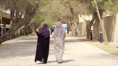 Riyad Parkı 'nda Yürüyen Kadınlar. Geleneksel abayalarda iki kadın, Suudi Arabistan 'ın Riyad kentindeki ağaçlarla çevrili gölgeli bir patikada yürüyor. Barışçıl dış mekan şehirdeki günlük yaşamı yakalıyor.