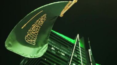 Ulusal Bayram kutlamalarında Suudi Bayrağı Gece Dalgalanıyor. Suudi Arabistan bayrağının yakın çekimleri Ulusal Bayram 'ı simgeleyen yeşil ışıklarla süslenmiş aydınlanmış bir kulenin önünde gururla dalgalanıyor.