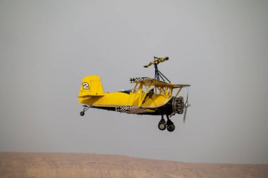 Wingwalker KSA Hava Gösterisi 'nde Suudi Çölü Havacılık Daring' in üstünde uçuyor. Korkusuz bir kanat yürüyüşçüsü, bir havacılık gösterisi sırasında çölde süzülen klasik sarı bir çift kanatlı uçağın tepesinde nefes kesici bir gösteri sergiliyor.