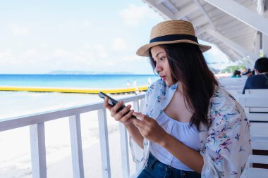Deniz kenarında akıllı telefon kullanan genç bir kadın. Mutlu turistlerin dinlenmesi ve cep telefonunu tropikal gündoğumu plajlarında sosyal ağ için kullanması iletişim teknolojisini kullanıyor. Seyahat kavramları ve teknoloji
