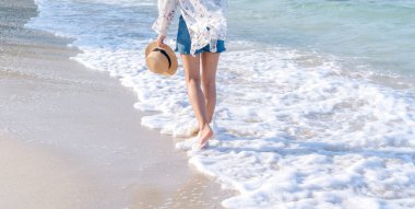 Yaz mevsiminde turkuaz renkli okyanusun yanında kumlu sahilde yürüyen bir kadının bacaklarının arka görüntüsü. Hasır şapkalı kadın turist güneşli bir günde egzotik doğaya seyahat ediyor..