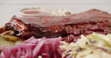 İçinde kirazlı chipotle soslu bebek kaburgaları olan büyük bir tabak. Sigara, sos, barbekü fasulyesi, lahana salatası, soğan turşusu, salatalık turşusu, izole edilmiş beyaz arka planda dönerek yavaş yavaş pişiyor.