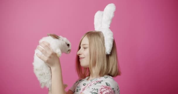 可愛いウサギちゃんと幸せな女の子 ピンクを背景にイースターのお祝いの間にウサギの耳が笑顔で愛らしい白いウサギと鼻に触れると幸せなブロンドの女の子の静的なショット ハッピーイースター — ストック動画