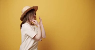 Boşluğu gösteren genç bir kadın. Beyaz tişörtlü ve hasır şapkalı, neşeli bir şekilde gülümseyen ve sarı arka plandaki boş alana bakan heyecanlı genç bir kadının statik çekimi..