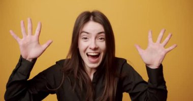 Heyecanlı genç bayan şaşırtıcı kamera. Şık siyah tişörtlü, elleriyle yüzünü örten, sarı arka planda haykıran ve gülen neşeli genç esmerin statik görüntüsü.