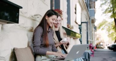 Laptop kullanan ve parmaklarına güvenen neşeli bir esmerin el kamerasıyla çekilmiş fotoğrafı. Elinde kahveyle bankta oturan ve şehir caddesinde uzaktan çalışan sarışın bir kız arkadaşın fotoğrafı.