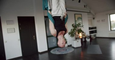 Adam havadaki lavabolarda baş aşağı asılı duruyor. Hafif stüdyodaki yoga seansında havadaki hamakta baş aşağı asılı duran güçlü bir erkeğin el kamerasıyla aşağı doğru dön ve vücudunu bük.