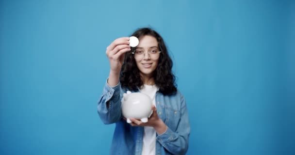 一个拿着比特币和钱箱的年轻女人身着休闲装 戴眼镜的年轻黑发女孩的静态照片 带有蓝色背景的比特币 — 图库视频影像