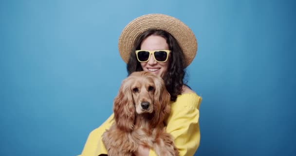 夏天带着狗的时髦女人 身着时髦太阳镜 头戴草帽的年轻女店主在蓝色背景下爱抚和亲吻英国可可卡犬的静态镜头 — 图库视频影像
