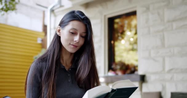 若いブルネットが路上で本を読んでいる 週末に街の通りで時間を過ごしながら本の中で面白い話を読んで長い暗い髪の若い女性のハンドヘルドショット — ストック動画