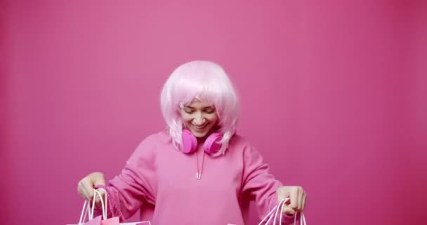 無線ヘッドフォンとピンクの髪を持つパーカーの陽気な若い女性はピンクの背景と販売中の笑みを浮かべて立っている間に購入で買い物袋を実証し チェック — ストック動画