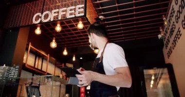 Beyaz tişörtlü, yoğun sakallı erkek barista siyah önlüklü ve tablet kullanan şeffaf yüz maskeli modern kahve dükkanında çalışırken kayıt yaptırıyor.