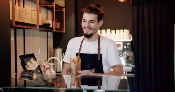 穿着白T恤 留着小胡子 面带微笑的年轻而积极的年轻男性咖啡师的低角度 在现代咖啡店的柜台上边吃小吃边看相机 — 图库视频影像