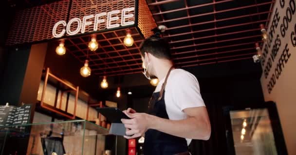 在现代咖啡店工作时 穿着白色T恤 留着浓密胡须的男性巴里斯塔 戴着黑色围裙 戴着透明的面罩 在工作时使用平板电脑 — 图库视频影像
