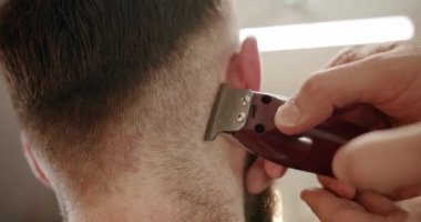 Yakışıklı adam elektrikli budayıcı ile yeni bir saç stili alıyor. Makyaj işiyle kapat. Modern berber dükkanında sakal ve bıyık bakımında genç hippi beyaz adam. Erkek saç stili. 