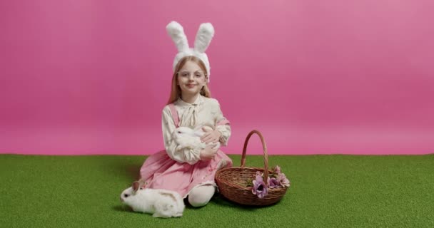 ピンクを背景にウィッカーバスケット近くの偽の芝生の上でひざまずきながら 笑顔でカメラを見て ふわふわのウサギを撫でるウサギの耳と眼鏡を持つ肯定的なブロンドの女の子 ハッピーイースター — ストック動画