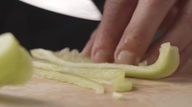 Genç aşçının önden görünüşü kesme tahtasındaki bıçakla ince dilimler halinde yeşil biber kesiyor. Bir parça biber kesen genç bir şefin ön görüntüsü. Sebzeler çok yakın..