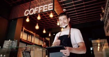 Beyaz tişörtlü, yoğun sakallı erkek barista siyah önlüklü ve tablet kullanan şeffaf yüz maskeli modern kahve dükkanında çalışırken kayıt yaptırıyor.
