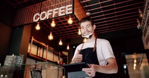 在现代咖啡店工作时 穿着白色T恤 留着浓密胡须的男性巴里斯塔 戴着黑色围裙 戴着透明的面罩 在工作时使用平板电脑 — 图库视频影像
