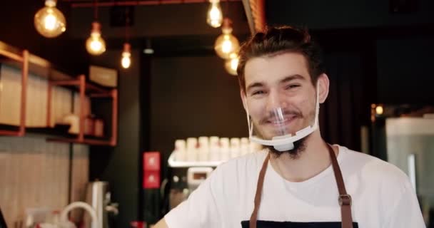 站在现代咖啡屋时 积极的年轻男性咖啡师笑着看着相机 在咖啡店工作时心满意足的年轻人笑了 店主站在酒吧里 — 图库视频影像