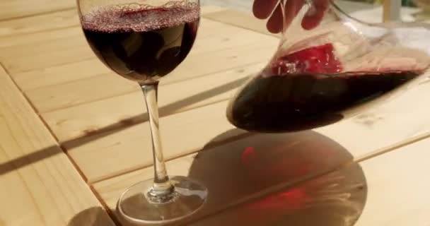 ソムリエは木製のテーブルの上でゆっくりと動きながら赤いワインをデカンタで振るときれいなガラスに注ぎ込まれます ワインはテーブルの上で光から屈折する 沈殿物や臭いのワインをきれいにする — ストック動画
