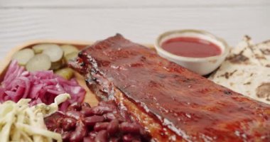 İçinde kirazlı chipotle soslu bebek kaburgaları olan büyük bir tabak. Sigara, sos, barbekü fasulyesi, lahana salatası, soğan turşusu, salatalık turşusu, izole edilmiş beyaz arka planda dönerek yavaş yavaş pişiyor.
