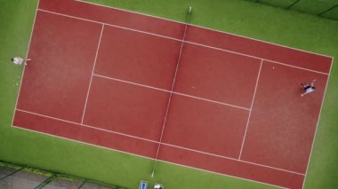 Arkadaşlar dışarıda halı kortlarında tenis oynarlar. Havadan, yukarıdan aşağıya, İHA atışı. Bir kadın profesyonel ve dinamik olarak yavaş çekimde tenis oynar. Eğlence için hafta sonu ve Pazar etkinliği.