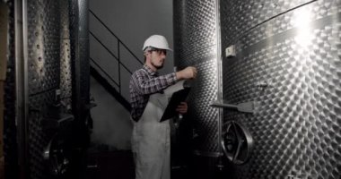 Erkek şarap imalatçısı, içeceklerin kalitesini içeride kontrol ediyor ve günlüğe tarihleri yazıyor. Şarap imalatçısı şarap fabrikasında damıtma ve fermantasyon için ekipman arıyor..