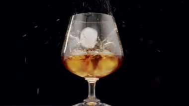 Luxury brandy. Glass with golden cognac on isolated black background with ice cube. Brandy, cognac, and whisky. Slow motion. Luxury Drink Whisky with smoke. The ice falls into the whiskey glass.