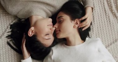 LGBT rights, Lesbian family. Low angle handheld shot of young lesbian couple smiling and touching hands gently while sitting on sofa against window in light living room at home.