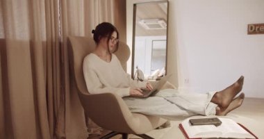 Güzel Genç Kadın, dizüstü bilgisayarda çalışırken sandalyede oturuyor. Defterde Kazak Giyen Şehvetli Kız. Çalışmalar, İnternet Sörfleri, Konforlu Daire 'de dinlenirken Sosyal Medya Kullanımı