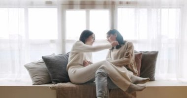 Low angle handheld shot of young lesbian couple smiling and touching hands gently while sitting on sofa against window in light living room at home. Lifestyle and relationships. LGBT concept