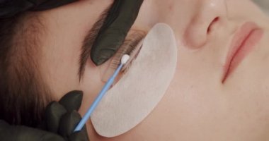 Crop cosmetologist doing eyelash extending procedure. From above closeup handheld shot of crop beautician using cotton swab to apply glue on eyelashes of woman during beauty session in salon.
