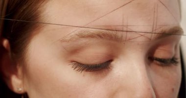 Crop cosmetician making creative makeup. Closeup handheld shot of crop makeup artist in latex gloves using thread to stamp lines on forehead of woman in salon.