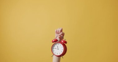 A hand raised up a red alarm clock on an isolated yellow background. The alarm clock serves as a symbol of the daily routine and the passage of time. importance of time management. 