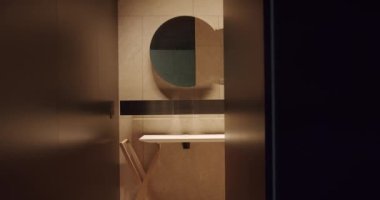 Modern bathroom behind opened door. Pan left view of geometric mirror and shelf installed on tiled wall behind opened door of contemporary bathroom in evening