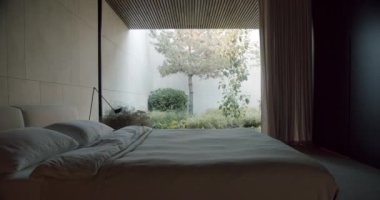 Modern bedroom with glass wall. Pan left view of comfortable bed located against glass wall with view of green garden in contemporary bedroom