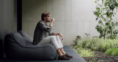 Man drinking tea on terrace. Static shot of relaxed male in casual clothes sitting on bean bag and sipping hot drink in morning on terrace