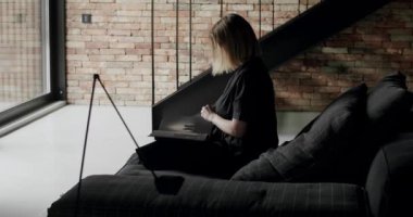 A woman is sitting on the black sofa and reading a book. Minimalist living room with bricks wall, large sliding windows and black furniture. Luxury Modern House Interior With Corner Sofa and Bookshelf