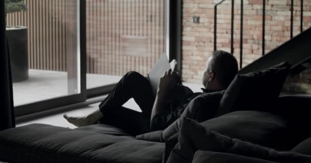 男が黒いソファに座って本を読んでいる 現代的なレンガの壁 大きなスライド窓と黒の家具とミニマリストのリビングルーム コーナーソファと本棚付きインテリア — ストック動画