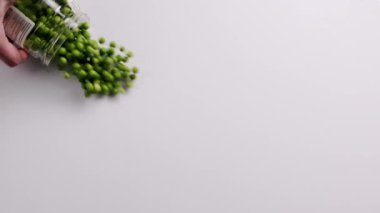 The mans hand rolls a jar full of green peas, which fills the surface. top view. Raw green peas in slow motion on white background.