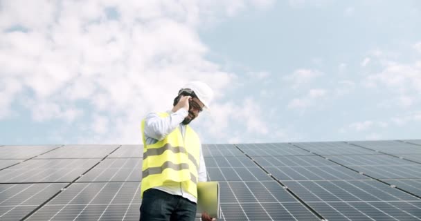 太陽光発電所のスマートフォンで話す男性エンジニア 太陽光発電所の晴れた日に太陽光発電パネルの近くを歩くと曇りの青空に対する電話に応答する制服を着た低角度の男 — ストック動画