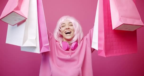 穿着连帽衫 头戴无线耳机 头戴粉色头发的快乐的年轻女性一边展示和检查购物袋 一边站在粉色背景下 一边在销售过程中微笑 — 图库视频影像