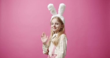 Static shot of cheerful girl say hello with bunny ears and glasses covering eyes and looking at camera with smile during holiday celebration against pink background. Happy Easter Concept.