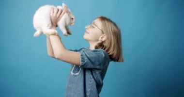 Happy girl with cute bunny. Static shot of glad blond girl with bunny ears smiling and touching noses with adorable white rabbit during Easter celebration against blue background. Happy Easter.