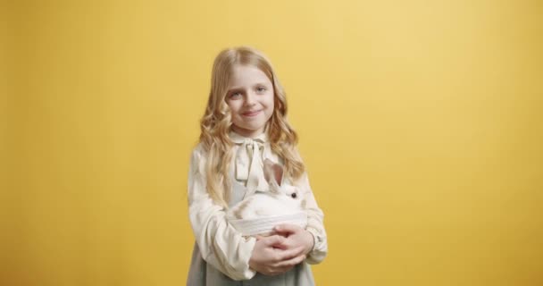 白いドレスの少女は 孤立した黄色の背景に彼女の手にウサギとバスケットを保持しています かわいいふわふわのウサギ 素敵な動物のコンセプト ハッピーイースターの日 バニー テキストメッセージのバナー — ストック動画