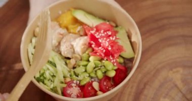 A woman hand serves Poke bowl with shrimp, rice, avocado, mango, edamame beans, cucumber on a wooden background. Healthy food, keto diet concept. Fresh fish and vegetables on diet lunch.