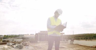 Male engineer in uniform and white helmet makes notes in notebook at work. Verification of works on the territory of the construction site. Industrial Building Construction Site. 